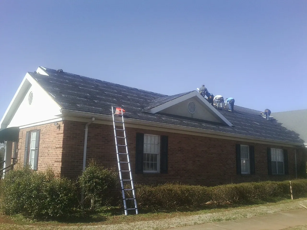 Crew tearing shingles off a brick ranch roof with a ladder set — Metal Roof Repair work by our crew in Avery