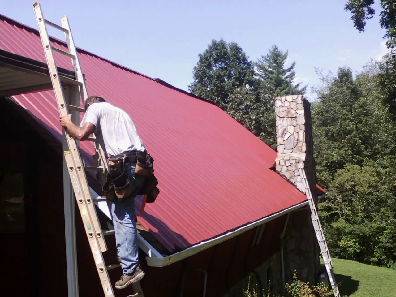 Roofer on a ladder beside a red metal roof and stone chimney — Soffit & Fascia Repair by Roofing Brothers in Avery